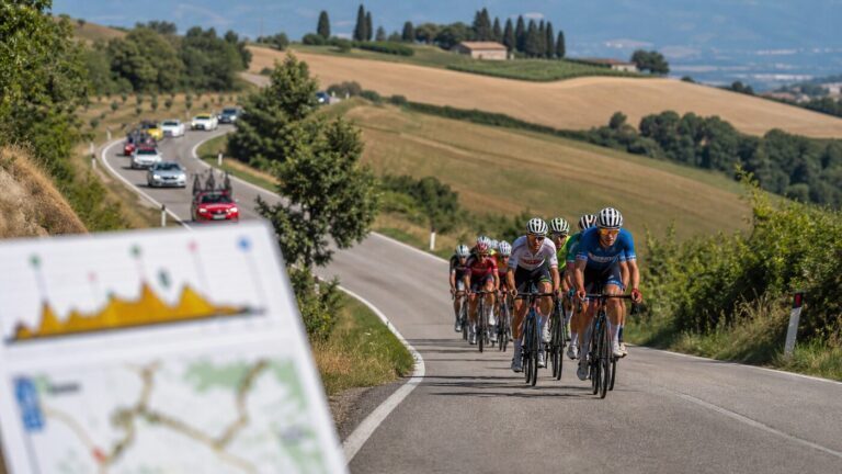 Gruppo di ciclisti su strada collinare con auto al seguito e profilo di tappa in primo piano